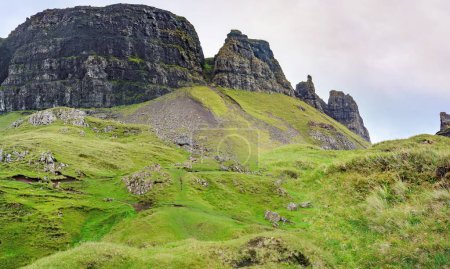 Photo for Sheer vertical sides of jagged dark rock faces,lining the Quraing escarpment,northernmost summit of the Trotternish,ancient landslide,mountain trail beneath covered with summer grass. - Royalty Free Image