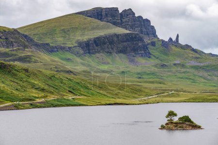 Téléchargez les photos : La Storr, au-delà du loch, au loin, un important point de repère Skye, falaises escarpées, petite île rocheuse saillante de l'eau, route de montagne à côté de l'eau du loch, jour d'été. - en image libre de droit