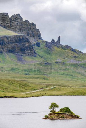 Foto de La Storr, más allá del lago, en la distancia, un prominente hito Skye, escarpadas caras de acantilado, pequeña isla de roca que sobresale del agua, camino de montaña junto al agua del lago, día de verano. - Imagen libre de derechos
