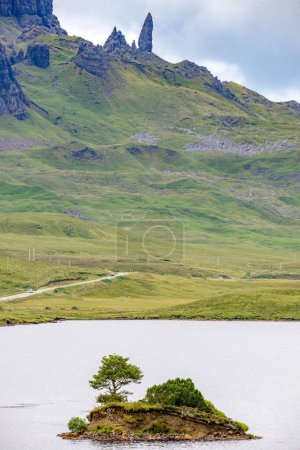 Foto de La Storr, más allá del lago, en la distancia, un prominente hito Skye, escarpadas caras de acantilado, pequeña isla de roca que sobresale del agua, camino de montaña junto al agua del lago, día de verano. - Imagen libre de derechos