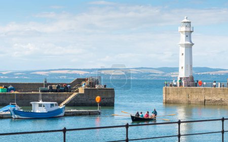 Téléchargez les photos : Édimbourg, Écosse - 30 juillet 2022 : Un petit bateau à rames en bois entre dans le petit port de pêche, transportant des passagers, flottant sur des eaux tranquilles et douces, par un après-midi chaud et ensoleillé en milieu d'été. - en image libre de droit