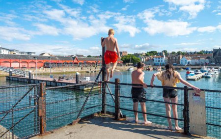 Téléchargez les photos : Édimbourg, Écosse-31 juillet 2022 : Habillés en maillot de bain par une chaude journée d'été, les enfants et les adolescents nagent et jouent dans les eaux calmes du port. - en image libre de droit