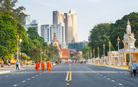Foto de Phnom Penh, Camboya-23 de diciembre de 2023: Vestidos con túnicas naranjas tradicionales, monjes, muchos con máscaras faciales, pasear por el Parque del Palacio Real, en una amplia carretera, cerrado al tráfico, en una tarde soleada. - Imagen libre de derechos