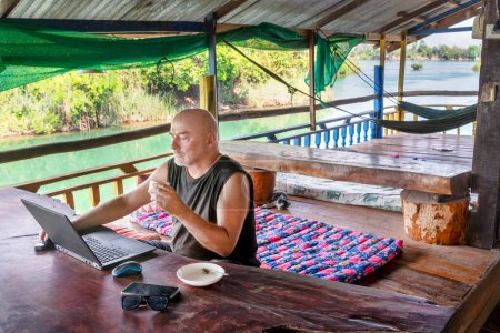 Foto de Trabajando remotamente en su computadora en Si Phan Don, un hombre que viaja por el mundo, se sienta en un balcón en una casa de huéspedes rural, con vistas al río Mekong, con una taza de café, en un entorno hermoso y relajado. - Imagen libre de derechos