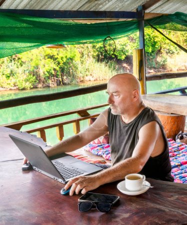Téléchargez les photos : Travaillant à distance sur son ordinateur à Si Phan Don, un homme voyageant à travers le monde, est assis sur un balcon dans une maison d'hôtes rurale, surplombant le Mékong, avec une tasse de café, dans un cadre magnifique et détendu. - en image libre de droit