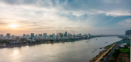 Photo for Rooftop view,looking across the river of the Cambodian capital city.Sun setting low in sky over Royal Palace and many high rise buildings.Beautiful gold sunlight reflecting from the river's surface. - Royalty Free Image