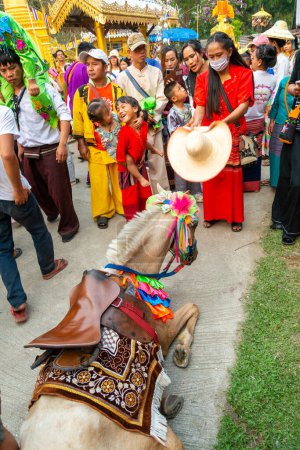 Photo for Pai,Northern Thailand-April 4th 2023: A crowd looks on,as a horse dressed in ceremonial garb collapses,and is helped by a woman, who uses her large hat as a fan, to try and cool the animal down. - Royalty Free Image