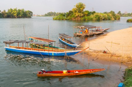 Foto de En el río Mekong, pequeños barcos de madera transportan a la gente hacia y desde las islas Nakasong y Don Som, en el punto del muelle de la playa de arena, ruta principal hacia la isla Don Det. - Imagen libre de derechos