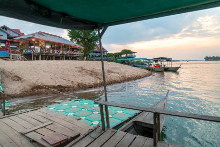 Foto de Al atardecer, en el río Mekong, pequeños barcos de madera que transportan a los viajeros hacia y desde las islas Nakasong y Don Som, en el punto del muelle de la playa de arena, puerta principal de entrada a la isla Don Det. - Imagen libre de derechos