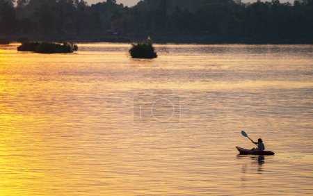Foto de Silueta de una persona en un Kayak, remando a lo largo de las tranquilas aguas del Mekong, en el archipiélago Si Phan Don, a través de rayos de luz dorada reflejados desde el sol poniente. - Imagen libre de derechos
