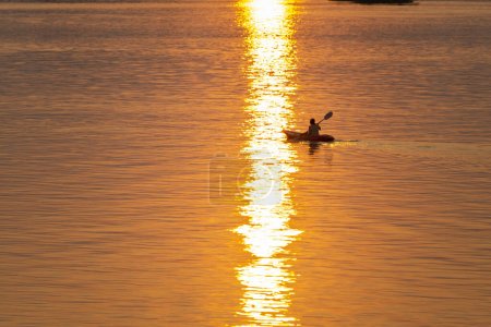 Foto de Silueta de una persona en un Kayak, remando a lo largo de las tranquilas aguas del Mekong, en el archipiélago Si Phan Don, a través de rayos de luz dorada reflejados desde el sol poniente. - Imagen libre de derechos