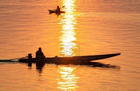 Foto de Siluetas de figuras humanas en pequeñas embarcaciones, a la deriva a través de las tranquilas y pacíficas aguas del Mekong, a través de rayos de luz dorada reflejados en el agua, desde el atardecer. - Imagen libre de derechos