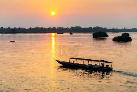 Foto de Silueta de embarcaciones fluviales, a la deriva a través de las tranquilas y tranquilas aguas del Mekong, a través de rayos de luz dorada reflejados en la superficie del agua del río, por el sol poniente. - Imagen libre de derechos