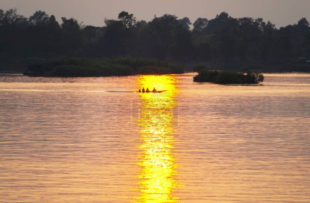 Foto de Siluetas de figuras humanas en una pequeña canoa, a la deriva a través de las tranquilas y tranquilas aguas del Mekong, a través de rayos de luz dorada reflejados en el agua, desde el sol poniente, en el sur de Laos. - Imagen libre de derechos