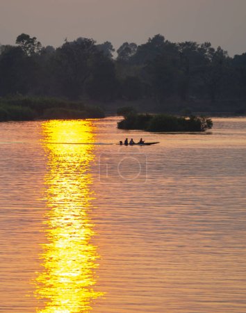 Foto de Siluetas de figuras humanas en una pequeña canoa, a la deriva a través de las tranquilas y tranquilas aguas del Mekong, a través de rayos de luz dorada reflejados en el agua, desde el sol poniente, en el sur de Laos. - Imagen libre de derechos