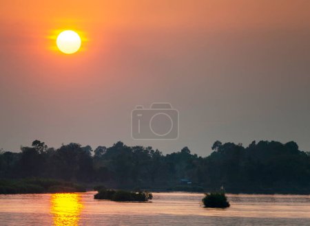 Foto de Si Phan Don.Silhouettes de pequeñas islas y árboles, a través de las tranquilas y tranquilas aguas del Mekong, el sol que se pone gordo colgando en el cielo.Luz dorada brillante reflejada en el agua, en el sur de Laos. - Imagen libre de derechos