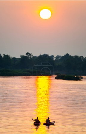 Foto de Siluetas de dos figuras humanas en Kayaks, a la deriva a través de las tranquilas y tranquilas aguas del Mekong, a través de rayos de luz dorada reflejados en el agua, desde el sol poniente, en el sur de Laos. - Imagen libre de derechos