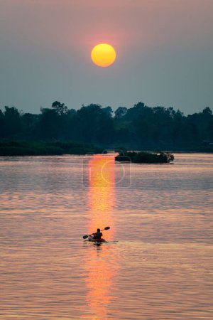 Foto de Silueta de una figura humana en un Kayak, a la deriva a través de las tranquilas y tranquilas aguas del Mekong, a través de rayos de luz dorada reflejados en la superficie del agua, desde el sol poniente, en el sur de Laos. - Imagen libre de derechos