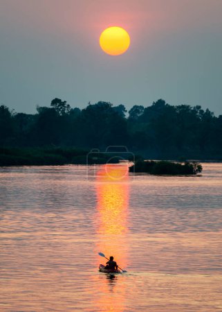 Foto de Silueta de una figura humana en un Kayak, a la deriva a través de las tranquilas y tranquilas aguas del Mekong, a través de rayos de luz dorada reflejados en la superficie del agua, desde el sol poniente, en el sur de Laos. - Imagen libre de derechos