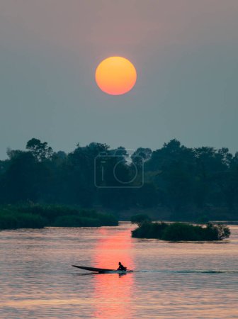 Foto de Silueta de figura humana en un pequeño y estrecho barco a motor, a la deriva a través de las tranquilas y tranquilas aguas del Mekong, a través de rayos de luz dorada reflejados en el agua, desde el sol poniente, en el sur de Laos. - Imagen libre de derechos