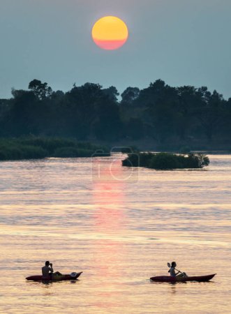 Photo for Silhouettes of two human figures in Kayaks,drifting across the calm,peaceful waters of the Mekong,through rays of golden light reflected on the water, from the setting sun,in southern Laos. - Royalty Free Image