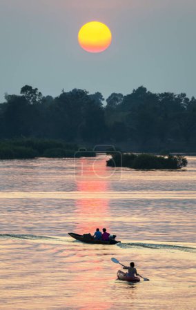 Foto de Siluetas de figuras humanas en pequeñas embarcaciones estrechas, a la deriva a través de las tranquilas y tranquilas aguas del Mekong, a través de rayos de luz dorada reflejados en el agua, desde el sol poniente, en el sur de Laos. - Imagen libre de derechos