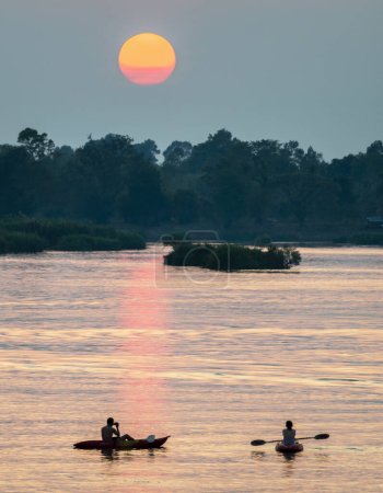 Foto de Siluetas de dos figuras humanas en Kayaks, a la deriva a través de las tranquilas y tranquilas aguas del Mekong, a través de rayos de luz dorada reflejados en el agua, desde el sol poniente, en el sur de Laos. - Imagen libre de derechos