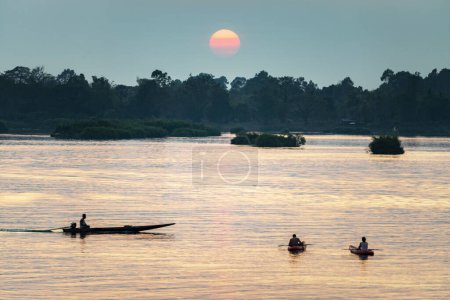 Foto de Siluetas de figuras humanas en pequeñas embarcaciones, a la deriva a través de las tranquilas y tranquilas aguas del Mekong, a través de rayos de luz dorada reflejados en el agua, desde el sol poniente, en el sur de Laos. - Imagen libre de derechos