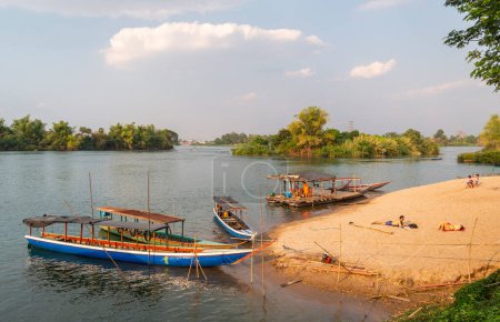 Foto de ,4000 Islands, Si Phan Don, Khong District, Laos-February 21 2023: Los viajeros se relajan en la diminuta playa del río Mekong, pequeños barcos de madera transportan a la gente hacia y desde las islas Nakasong y Don Som hasta aquí.. - Imagen libre de derechos