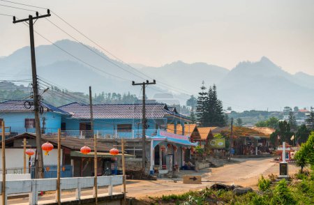 A narrow,dusty road,leading from Pai bridge,lined with small shops and small,colorful residential buildings,with dramatic mountain scenery in the background.