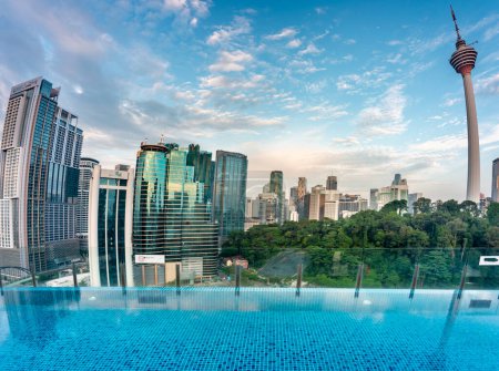 Superbe vue sur le toit de la ville de KL et Menara Kuala Lumpur, près du coucher de soleil avec bar sur le toit bleu piscine à débordement au premier plan et ciel bleu parsemé de nuages, gratte-ciel et arbres dans le parc KLCC.