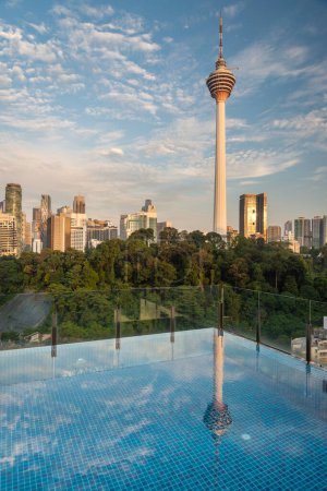 Atemberaubender Blick auf die Stadt KL und Menara Kuala Lumpur, bei Sonnenuntergang mit blauem Infinity-Pool auf der Dachterrasse und blauem Himmel mit Wolken. Moderne Wolkenkratzer und Bäume in einem Park.