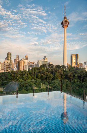 Atemberaubender Blick auf die Stadt KL und Menara Kuala Lumpur, bei Sonnenuntergang mit blauem Infinity-Pool auf der Dachterrasse und blauem Himmel mit Wolken. Moderne Wolkenkratzer und Bäume in einem Park.