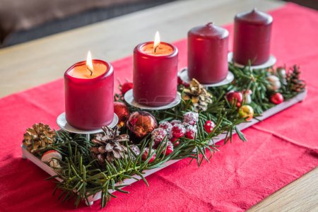 Photo for Burning red advent candles with decoration on a wooden table in living room on red fabric cloth at Christmas time, Germany - Royalty Free Image
