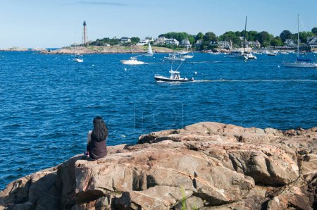 A woman sitting on rocks at Galehead overlooking the Marblehead Harbor and lighthouse in massachusetts.