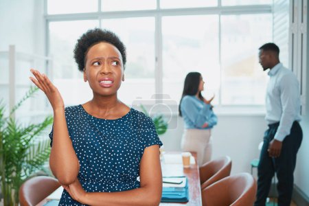 Foto de Mujer negra se ve molesta mientras sus colegas hablan a sus espaldas, drama de oficina. Foto de alta calidad - Imagen libre de derechos