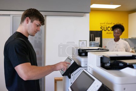 Téléchargez les photos : Un jeune homme utilise une photocopieuse dans un espace de travail partagé avec un collègue. Photo de haute qualité - en image libre de droit
