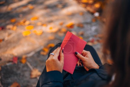 Photo for A woman opens a red envelope while sitting on a bench in a city park. Red envelope without inscriptions on a background of autumn foliage. High quality photo - Royalty Free Image
