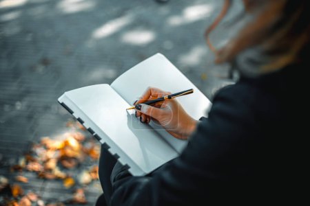 Photo for Young woman writing in a diary in an autumn park. Closeup image of a woman writing on a blank notebook. Woman working outdoors. High quality photo. - Royalty Free Image