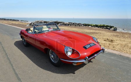 Photo for FELIXSTOWE, SUFFOLK, ENGLAND -  MAY 01, 2016: Classic  Red E-Type Jaguar Car parked on seafront promenade beach and sea in background. - Royalty Free Image