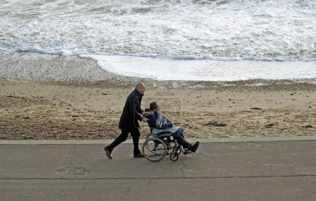 Photo for FELIXSTOWE, SUFFOLK, ENGLAND - DECEMBER 28, 2023:  Woman in wheelchair with foot fracture boot on being pushed alone seafront promenade by man  beach and sea in background. - Royalty Free Image