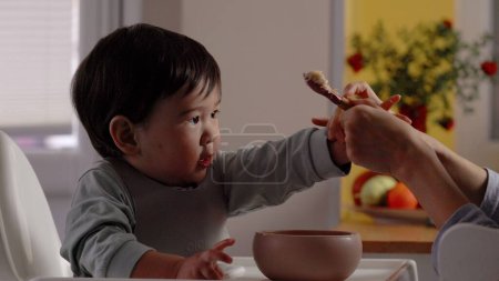 Téléchargez les photos : Asiatique bébé essaie de prendre une cuillère de sa mère pour manger sur son propre. mère prend cuillère et bol de nourriture du bébé. Images 4k - en image libre de droit