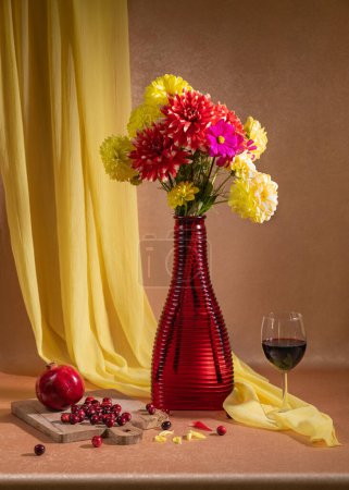 Photo for There are many bright flowers in a red vase, a glass of red wine stands nearby, red berries and pomegranates lie. Orange background and yellow handkerchief. Still life - Royalty Free Image