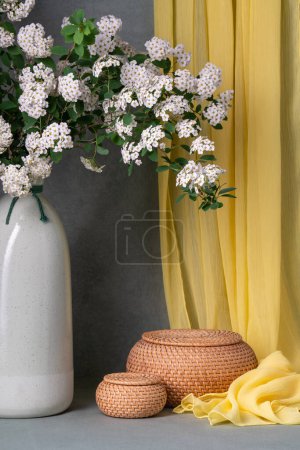 Still life with white flowers, spirea. Sprigs of white flowers in a brown vase on a gray background, next to two round baskets. Lots of places for text. Postcard, wishes 