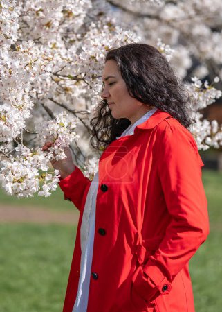 romantisches Bild einer stilvollen Frau in rotem Mantel, in weißer Bluse. Positive Stimmung. Ein nettes Mädchen hält sanft einen Zweig weißen Sakura und blickt lächelnd auf die Blumen.