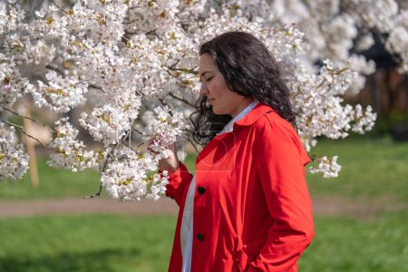 imagen romántica de una mujer con estilo en un abrigo rojo, en una blusa blanca. Humor positivo. Una linda chica suavemente sostiene una rama de sakura blanca y mira las flores, sonriendo.