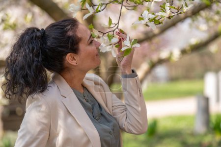romantisches Bild einer stilvollen Frau in heller Jacke. Positive Frühlingsstimmung. Ein nettes Mädchen hält sanft einen weißen Sakura-Zweig in der Hand und blickt lächelnd auf die Blumen