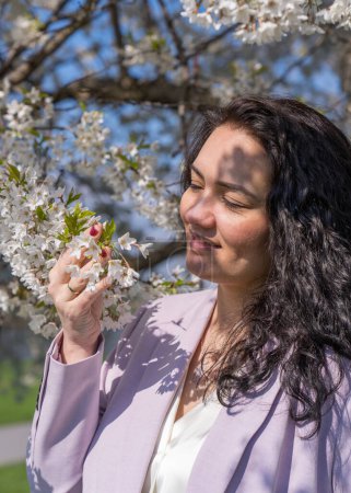 romantisches Bild einer stilvollen Frau in heller Jacke. Positive Frühlingsstimmung. Ein nettes Mädchen hält sanft einen weißen Sakura-Zweig in der Hand und blickt lächelnd auf die Blumen.