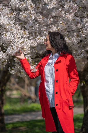 romantic image of a stylish woman in a red coat, in a white blouse. Positive mood. A cute girl gently holds a branch of white sakura and looks at the flowers, smiling.