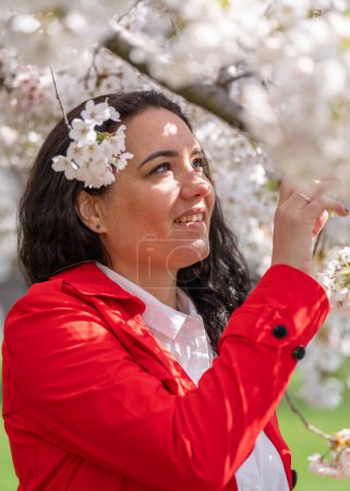 romantisches Bild einer stilvollen Frau in rotem Mantel, in weißer Bluse. Positive Stimmung. Ein nettes Mädchen hält sanft einen Zweig weißen Sakura und blickt lächelnd auf die Blumen